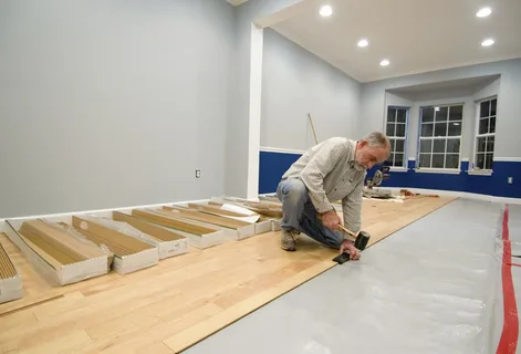 Contractor installing light-colored engineered wood flooring planks over an existing floor in a large commercial or residential space using a rubber mallet.